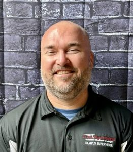Smiling man with a beard wearing a black shirt with "St. Vrain Valley Schools" logo, standing against a brick backdrop.