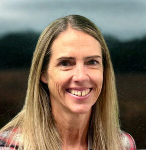 Smiling woman with long, straight hair, wearing a plaid shirt, against a blurred outdoor background.