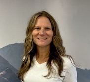 Smiling woman with long wavy hair, wearing a white shirt, standing in front of a mountain backdrop.