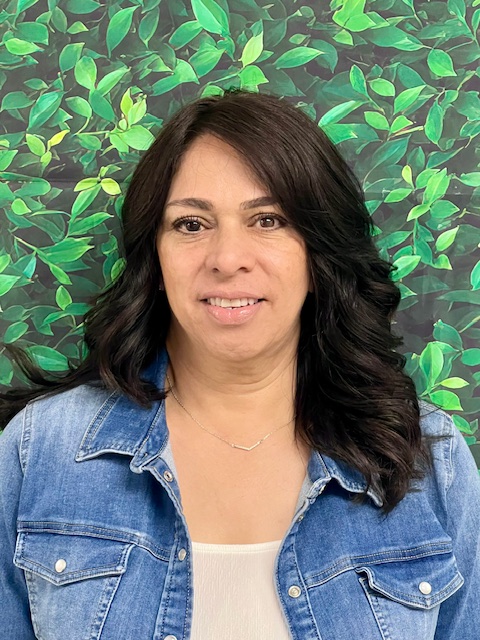 Woman with long, dark hair wearing a denim jacket, smiling in front of a green leafy background.