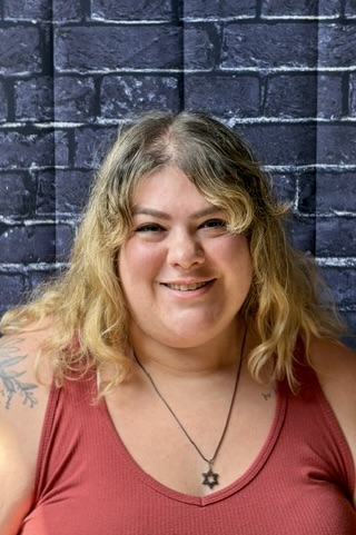 Smiling woman with wavy blonde hair, wearing a red tank top and a star necklace, against a dark brick background.