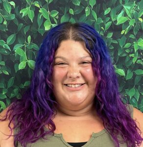 A smiling person with long, curly purple and blue hair stands in front of a leafy green background.