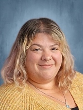A smiling person with wavy, shoulder-length hair, wearing a yellow sweater, against a blue background.
