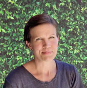 A woman with short hair smiles softly, set against a background of lush green leaves.