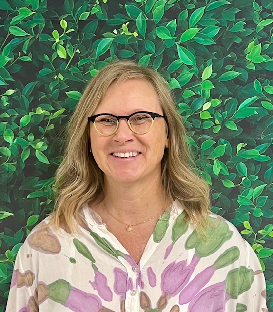 Smiling woman with wavy blonde hair wearing glasses, in front of a green leafy background.