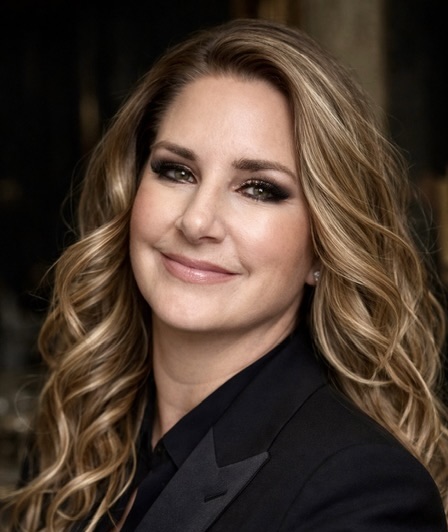 A woman with long, wavy hair smiles warmly, wearing a black suit jacket against a softly blurred background.