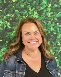 Smiling woman with brown hair in a denim jacket, standing in front of a leafy green backdrop.