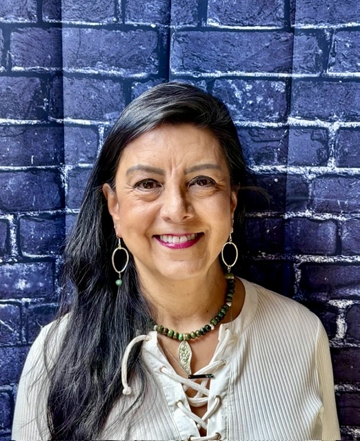 A woman with long dark hair smiles, wearing a beige top and jewelry, against a dark brick wall background.