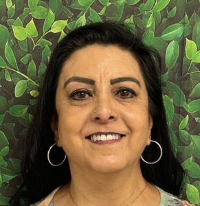 A woman with long black hair and hoop earrings smiles against a leafy green background.