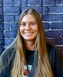 A smiling young woman with long hair poses in front of a dark brick wall, wearing a black shirt.