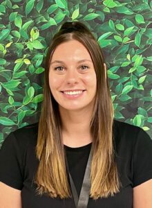 Smiling woman with long brown hair stands against a leafy green background, wearing a black shirt.