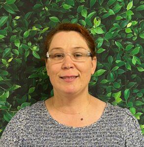 A woman with glasses smiles in front of a leafy green background, wearing a blue patterned top.