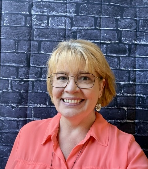 Smiling woman with short blonde hair and glasses, wearing a bright pink shirt, against a dark brick background.