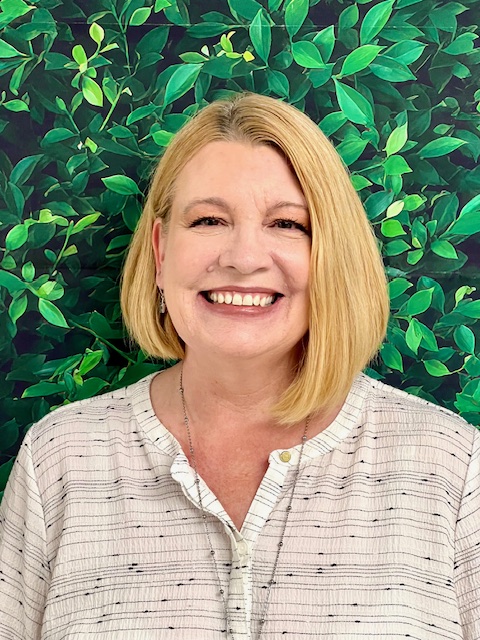 Smiling woman with blonde hair, wearing a light blouse, against a background of green leaves.