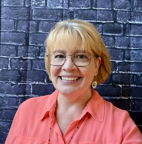 Smiling woman with short blonde hair, wearing glasses and a coral shirt, stands in front of a dark brick wall.