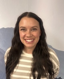 Smiling woman with long, wavy dark hair, wearing a striped shirt, standing in front of a light background.