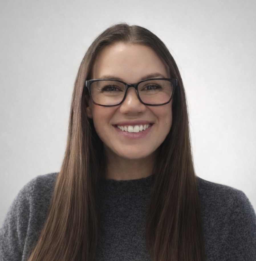 Smiling woman with long brown hair and glasses, wearing a gray sweater against a light background.
