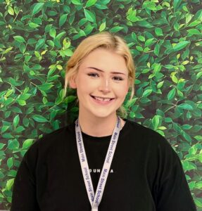 Smiling young woman with blonde hair, wearing a black shirt and a school lanyard, against a leafy green background.