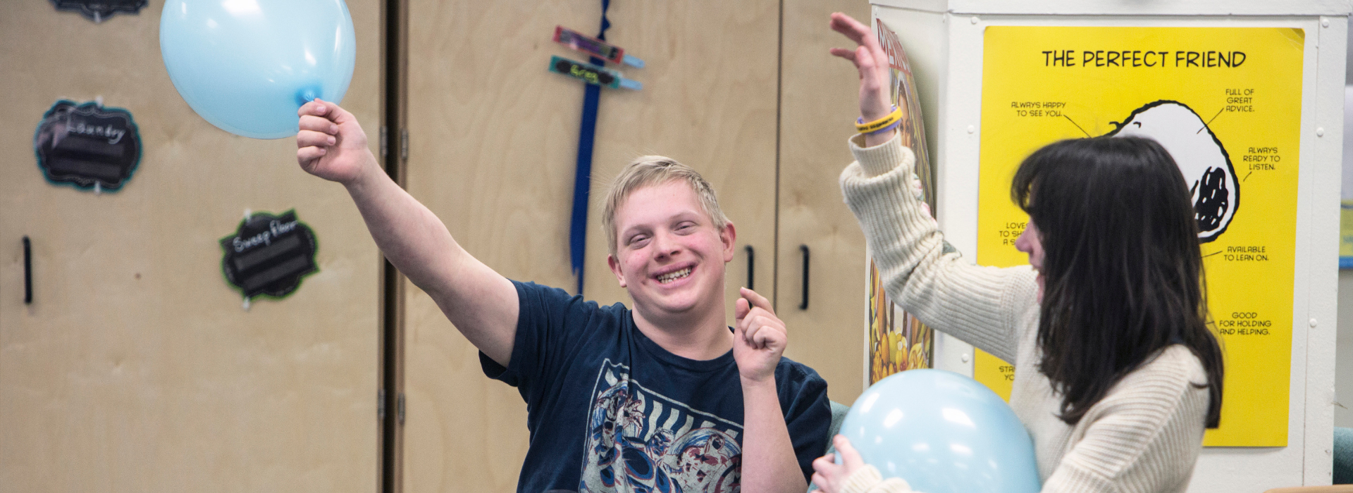 Dos personas celebran con globos azules en un aula llena de alegría, sonriendo y disfrutando del momento.